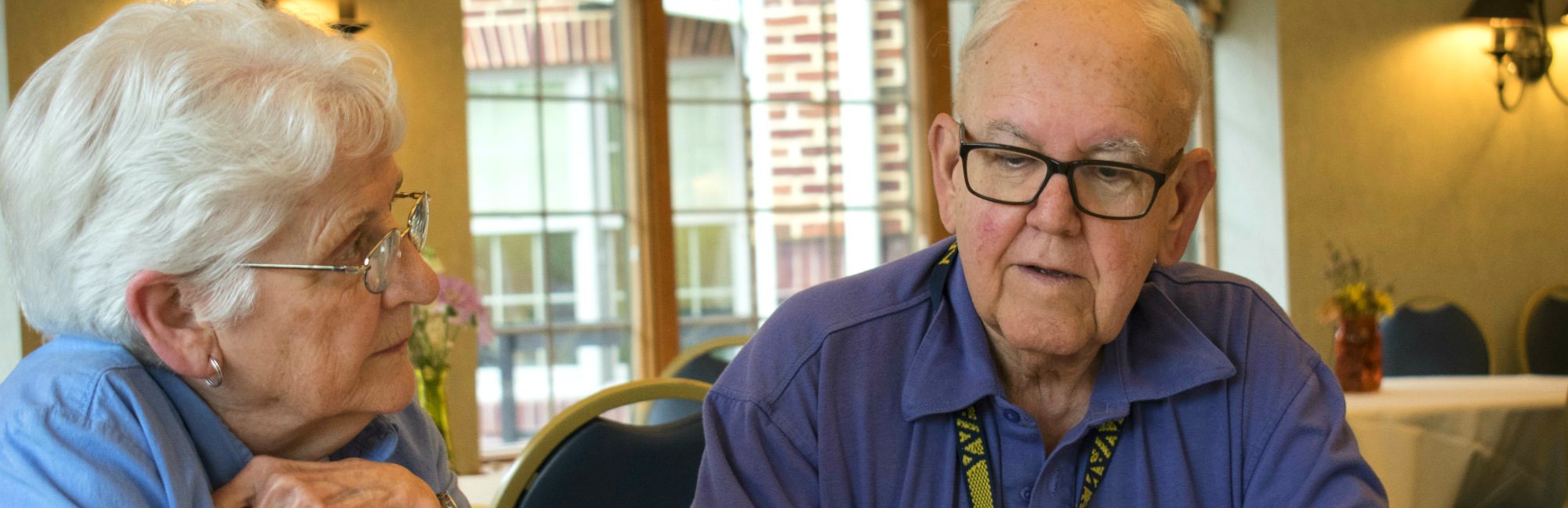 an elderly man wearing glasses and a lanyard sitting next to an older woman