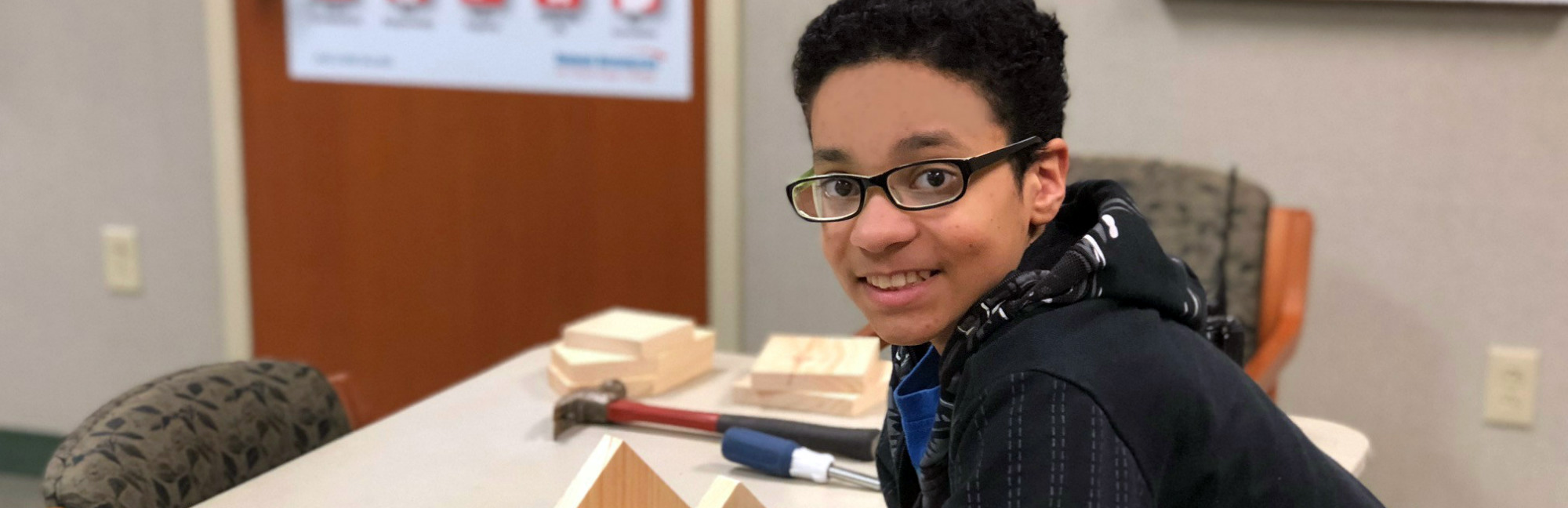 a young boy wearing glasses sits at a table with a hammer and a wrench