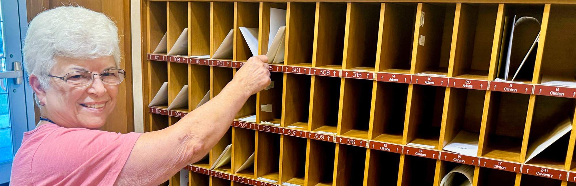 an elderly woman in a pink shirt is putting a piece of paper in a mailbox